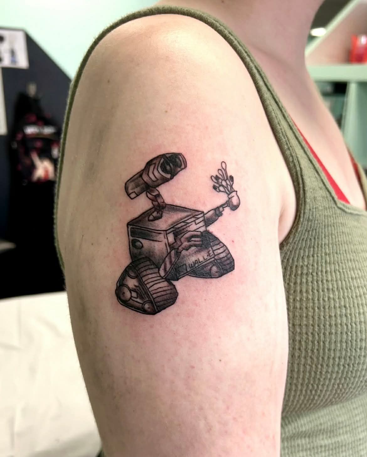 Tattoo of WALL-E holding a plant on a person's upper arm. Black and white, detailed. Person wears a green tank top.