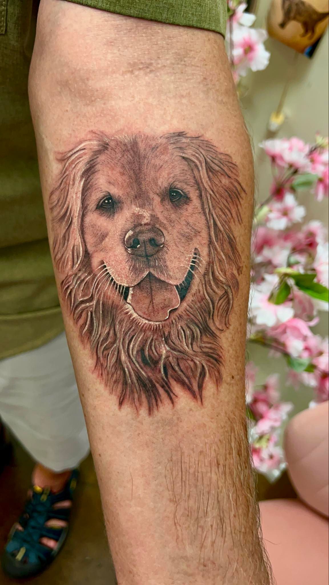 Dog portrait tattoo on a person's arm; tan fur, open mouth smile, set in a room with flowers.