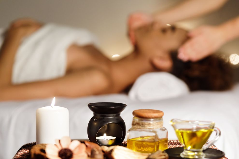 A reed diffuser, two lit candles, stacked grey stones, and white plumeria flowers on a wooden table in a massage setting.