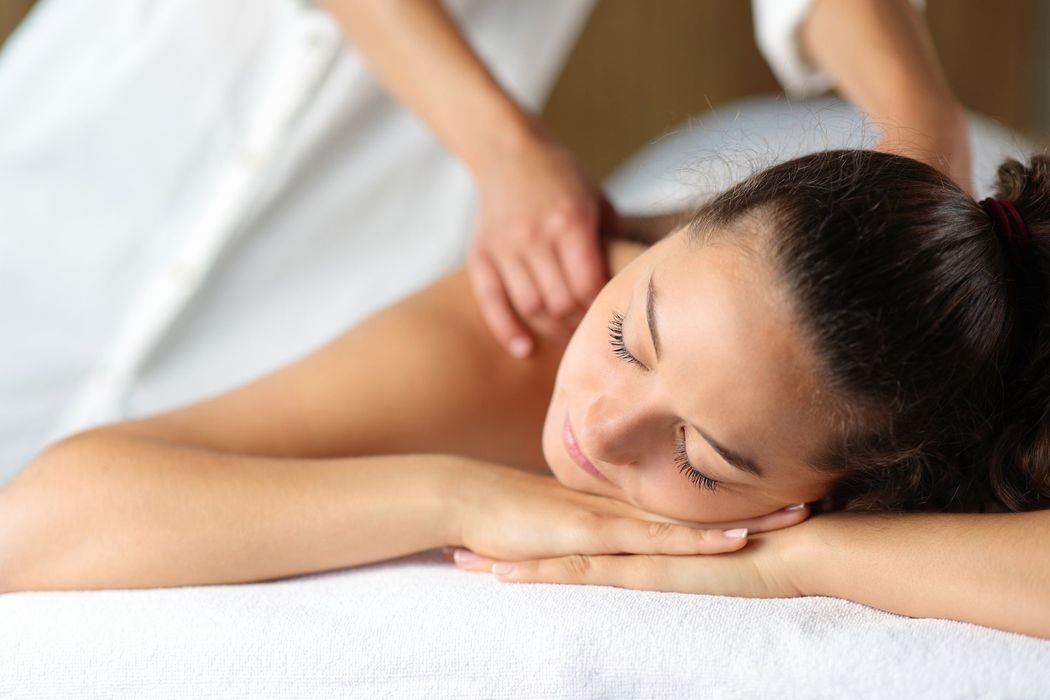 A person lying on a white surface with their eyes closed while receiving a massage on their shoulders.