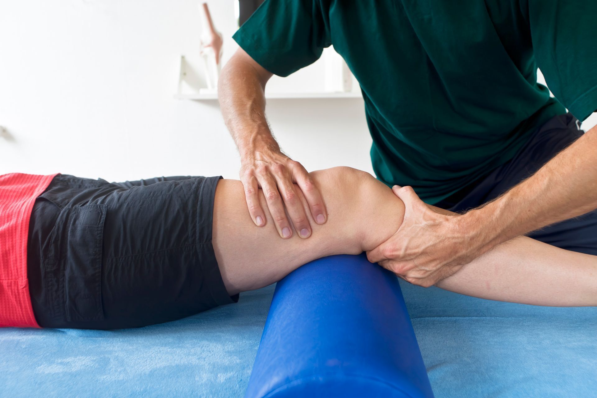 A physical therapist performs knee joint mobilization on a patient's leg, supported by a blue foam roller on a blue table. A physical therapist performs knee joint mobilization on a patient's leg, supported by a blue foam roller on a blue table.