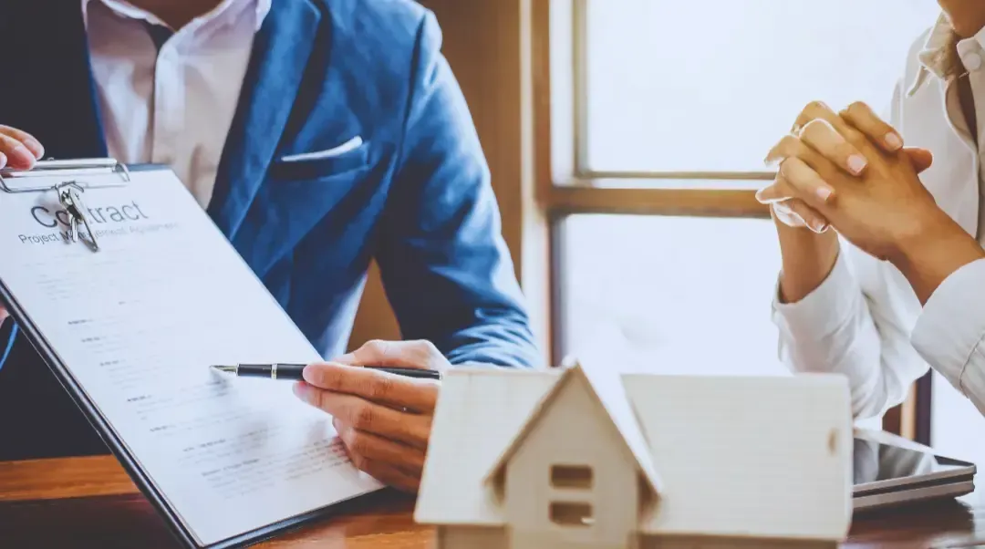 A man and a woman are sitting at a table with a model house and a clipboard.