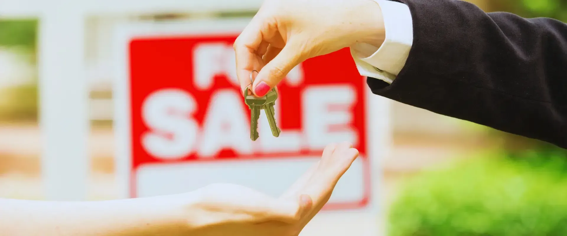 A person is handing a set of keys to another person in front of a for sale sign.