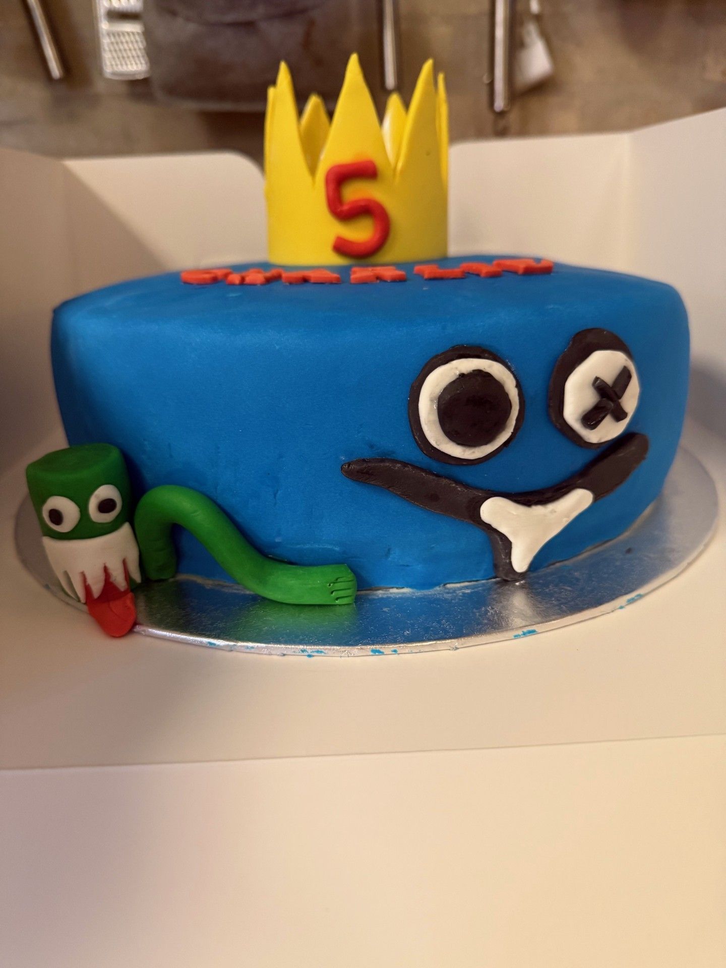 Blå fondantkage med et tegneserieansigt, en gul krone og en lille grøn figur.