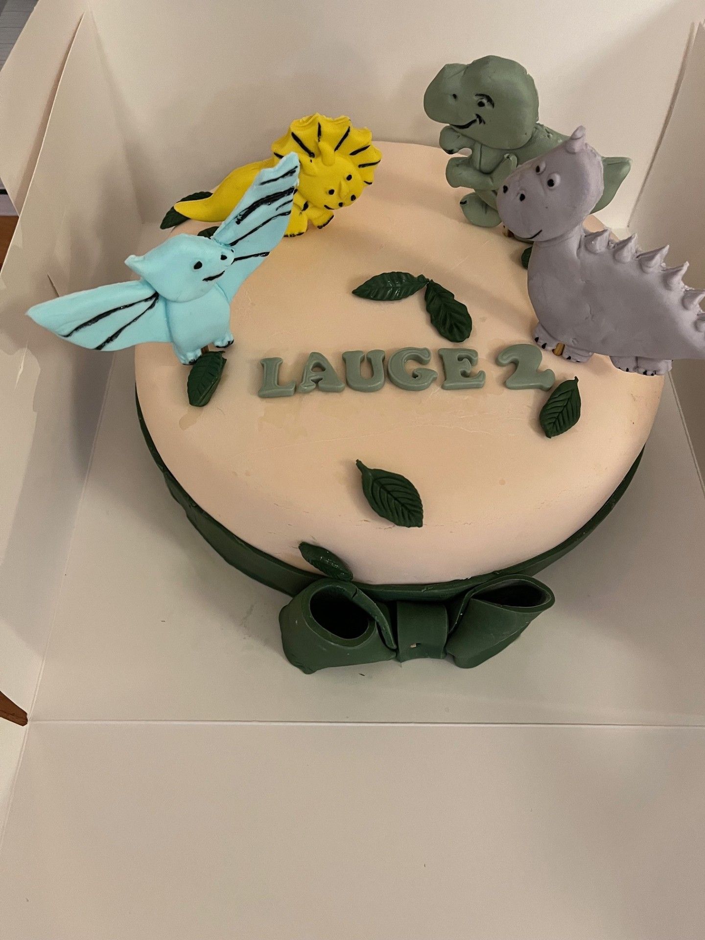 Fødselsdagskage med dinosaurtema i en æske med fondantpynt, inklusive en sløjfe og navnet
