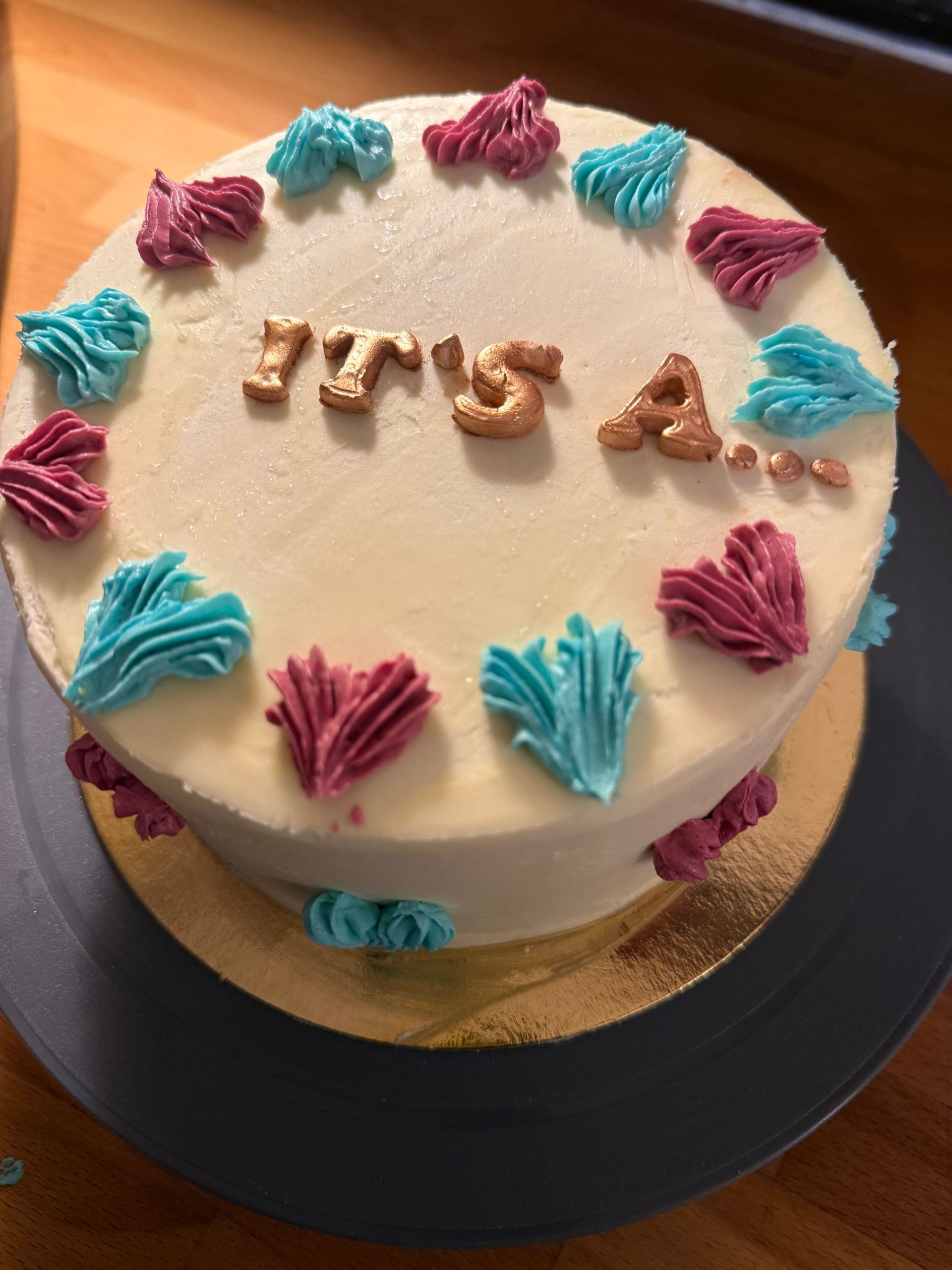 Rund kage med lyserød og blå frosting og guld “Det er”-topper på en sort fod