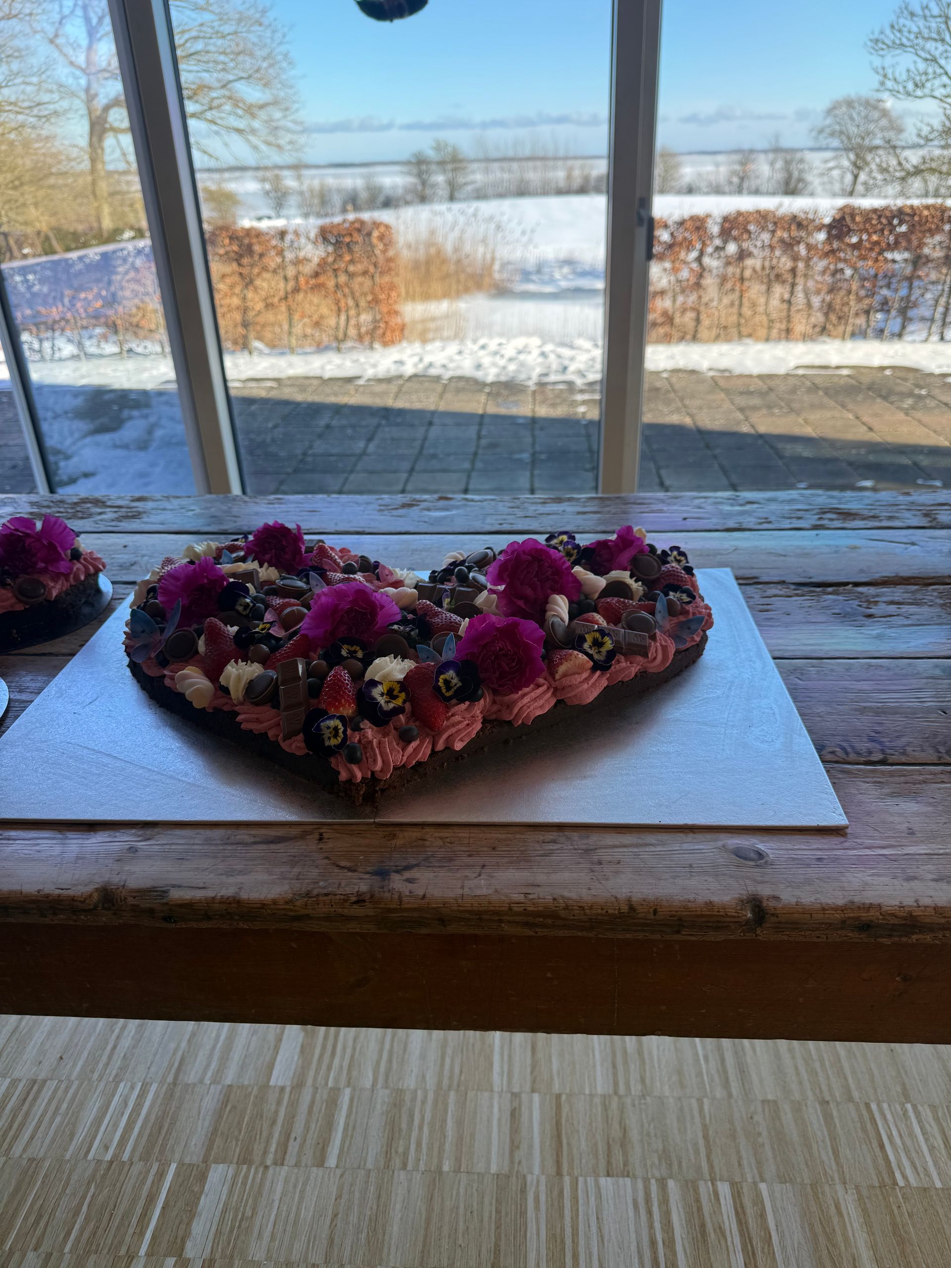Hjerteformet blomsterarrangement på en vindueskarm med udsigt over et snedækket landskab.