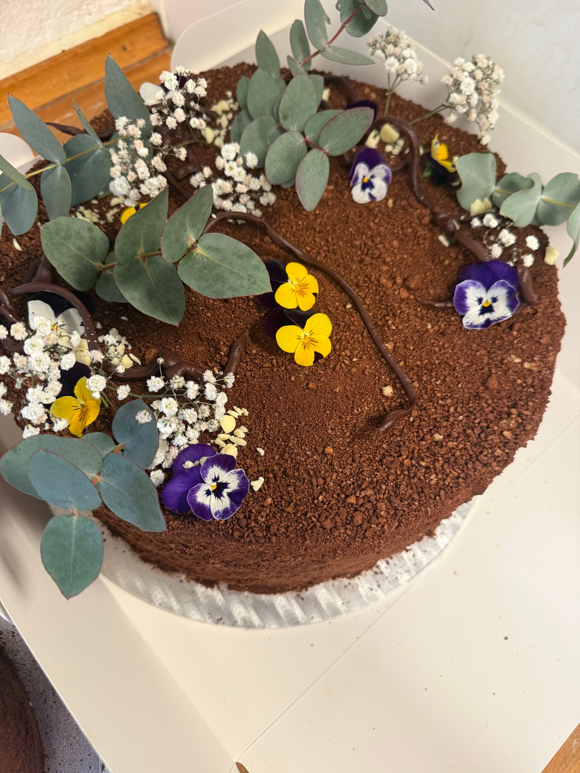 Rund chokoladekage med hvid frosting, grønne blade og små gule og lilla blomster på toppen