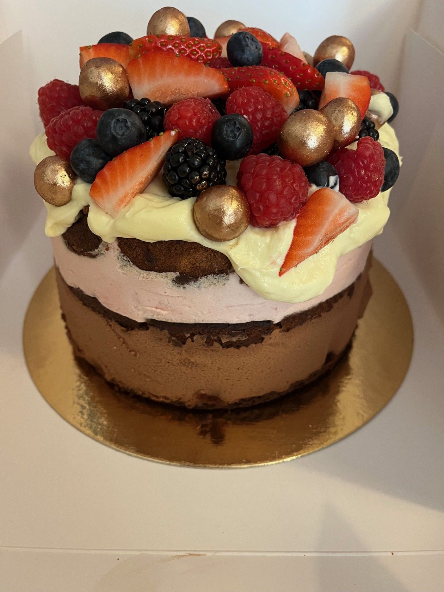 Kage med chokolade og lyserøde lag, toppet med fløde, bær og guldfarvede kugler.