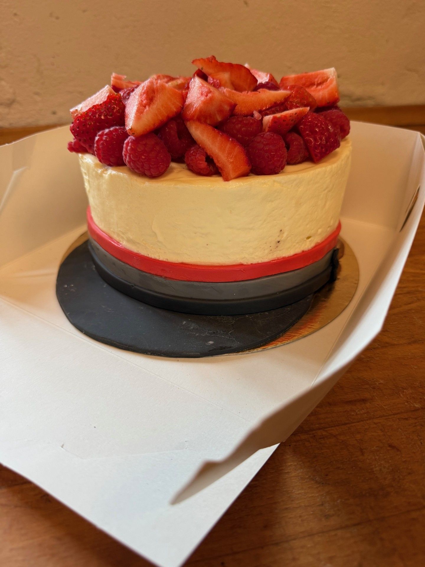 Kage med cremeglasur, lagdelt med rødt og gråt bånd, toppet med jordbær og hindbær, i en hvid æske.