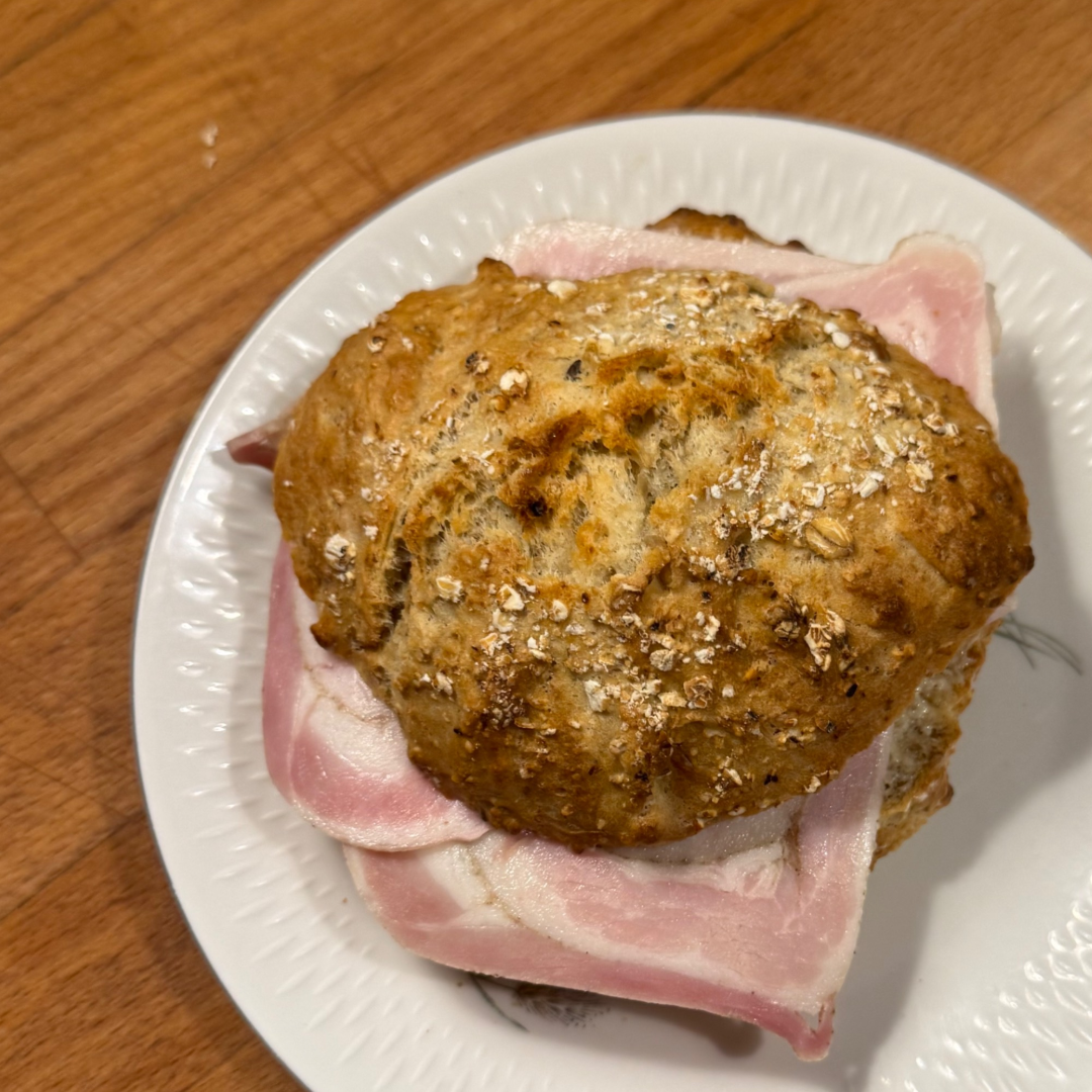 En sandwich af en brunet bolle på en hvid paptallerken med lyserøde skinkeskiver nedenunder.