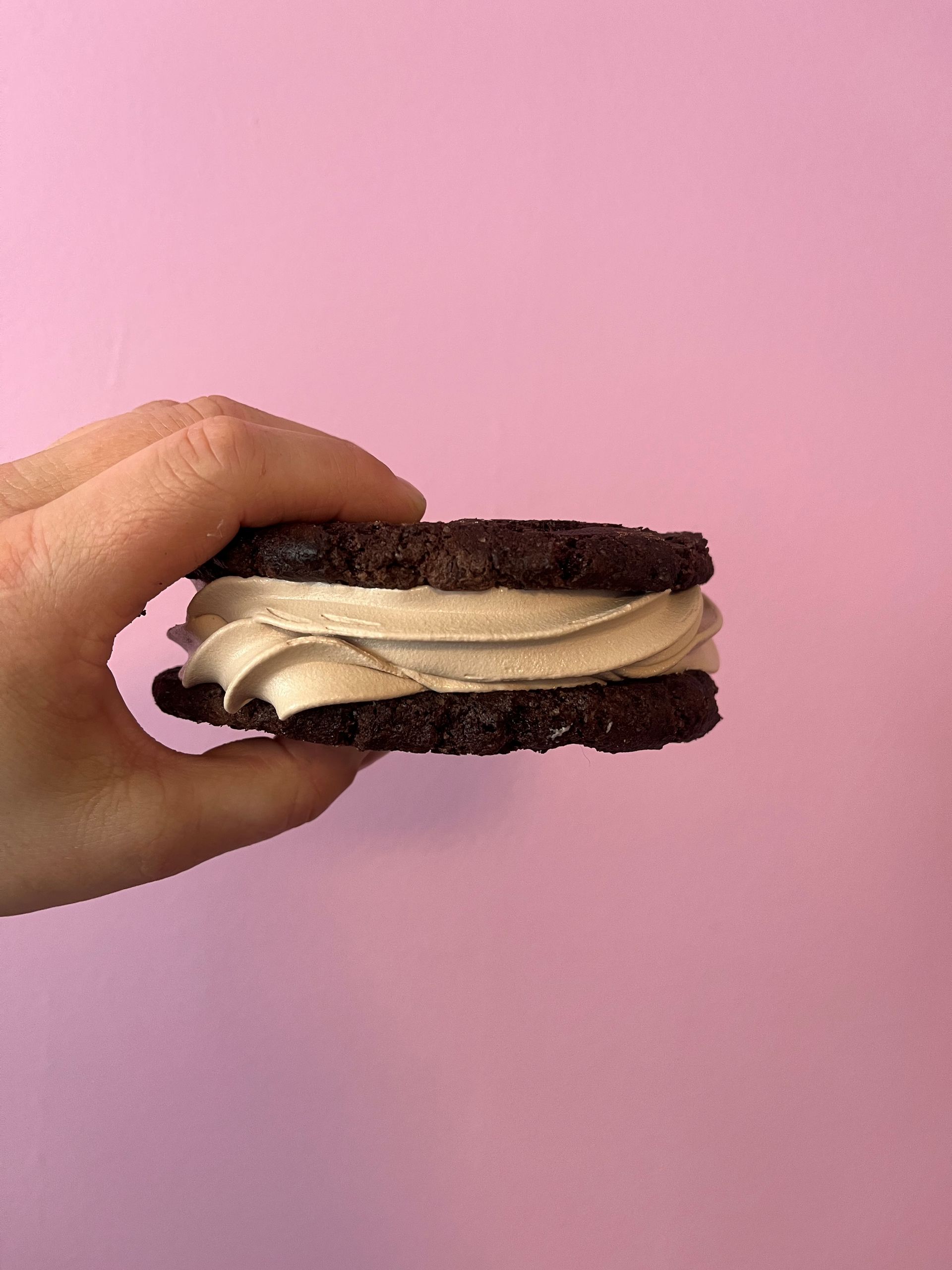 Hånd, der holder en chokoladekagesandwich med lyst fyld mod en lyserød baggrund.