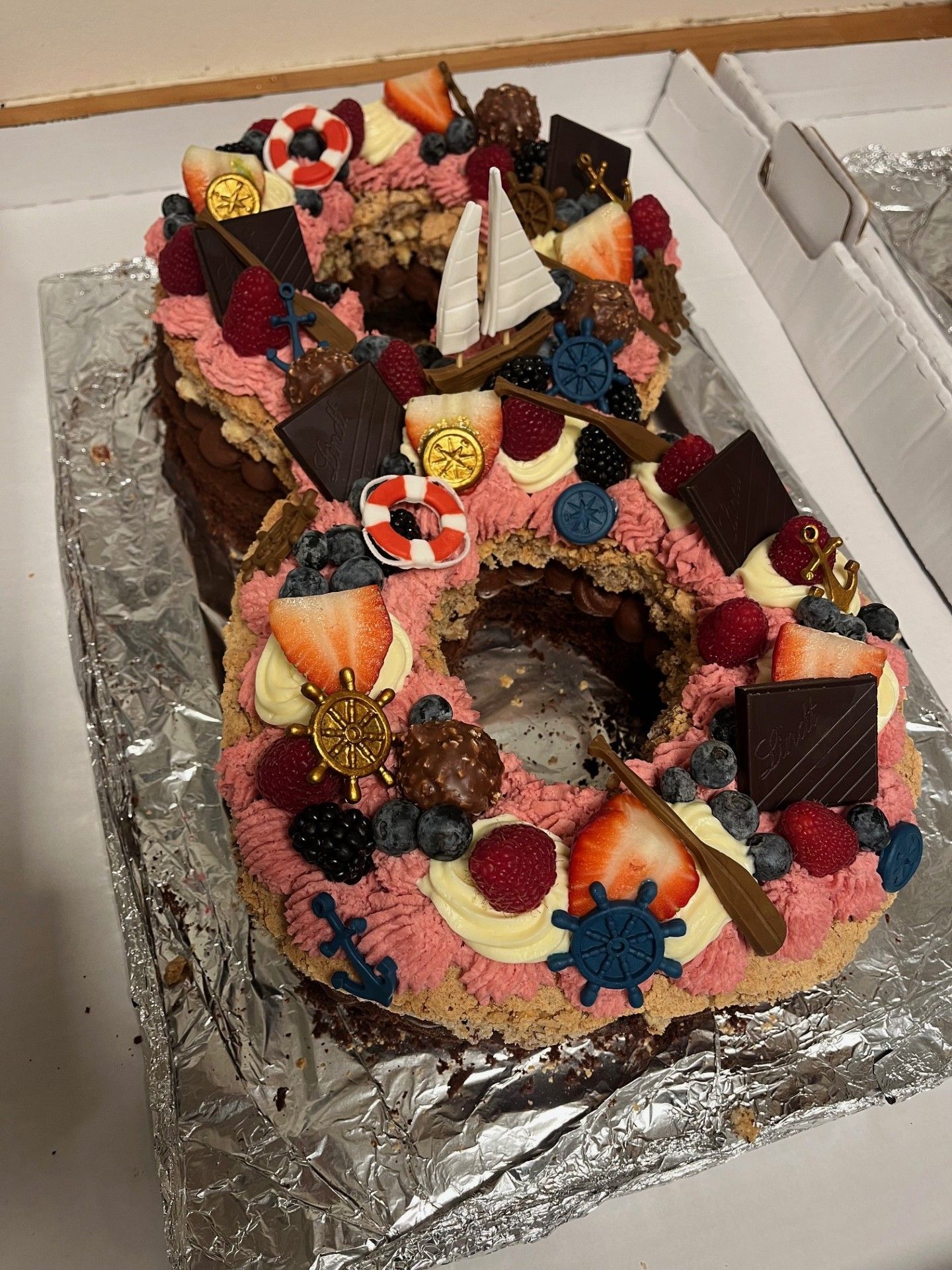 Talformet kage dekoreret med lyserød frosting, bær, chokolade og nautisk tema ornamenter.