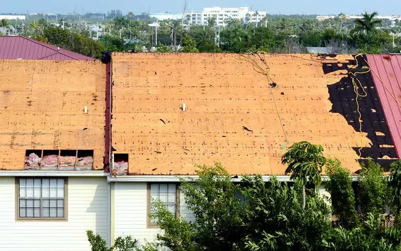 Damaged orange roof with exposed wood and missing sections. White building with windows, green trees in the foreground.