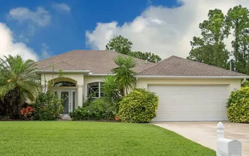 A single-story beige house with a white garage door, set in a green lawn under a blue sky.