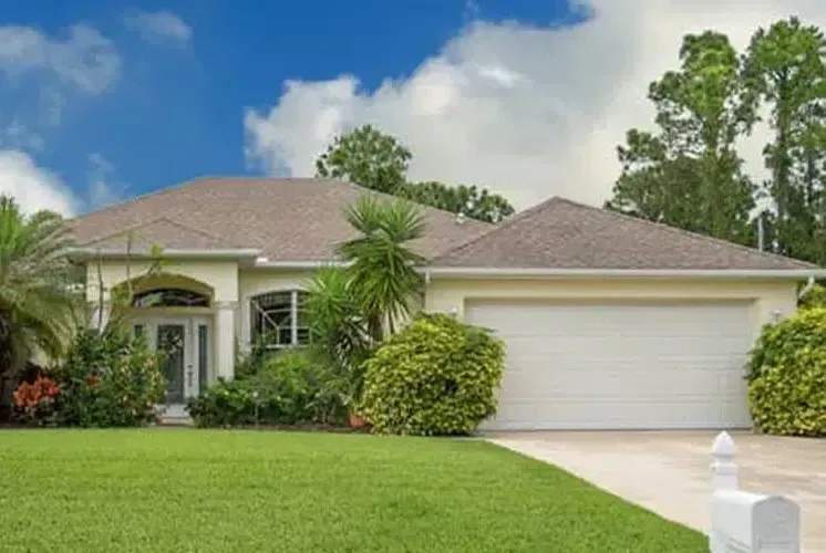 Single-story beige house with a two-car garage, lush green lawn, and blue sky.