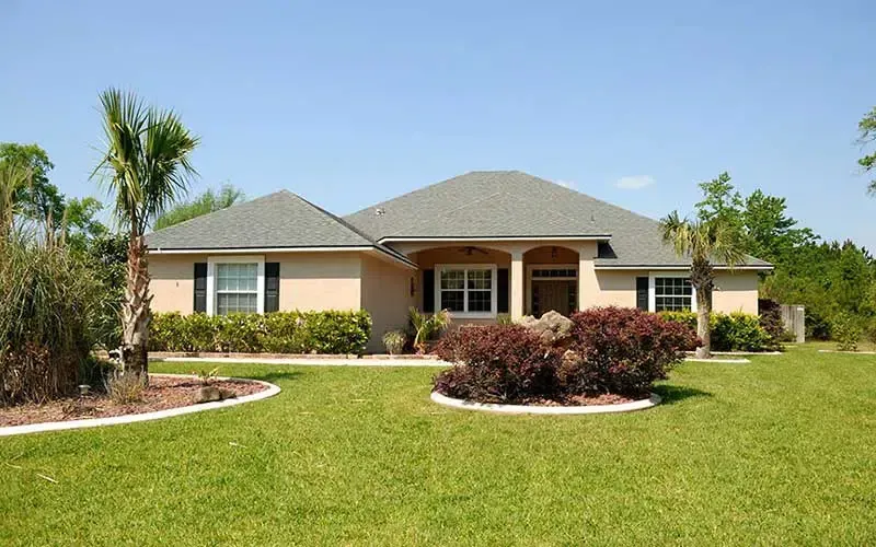 Beige ranch-style house with dark shutters, green lawn, and palm trees under a blue sky.