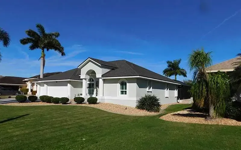 A light gray house with a dark roof and blue sky, palm trees in yard.