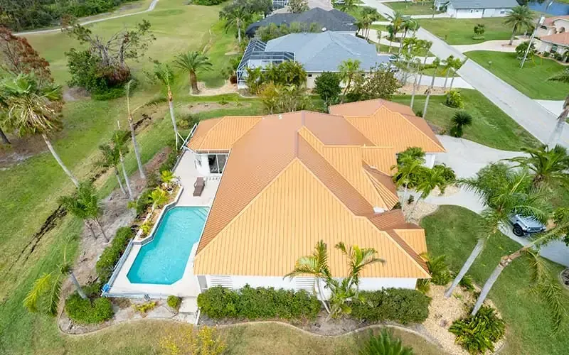 Aerial view of a white house with an orange roof, pool, and palm trees. Located in a grassy neighborhood.