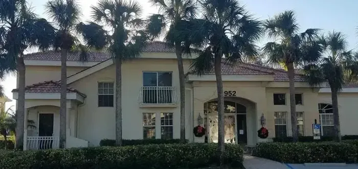 A beige two-story building with red-tiled roof, palm trees, and a front entrance with a sign.