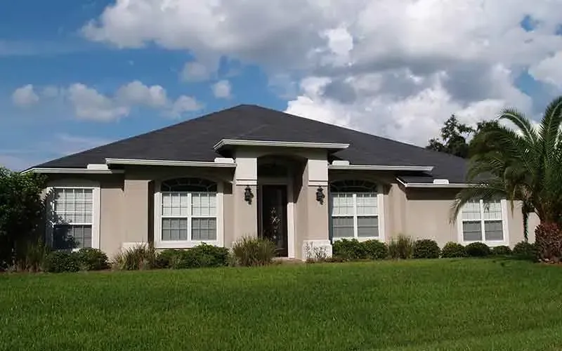 Tan house with black roof, white trim, windows, and green lawn under a blue sky with clouds.