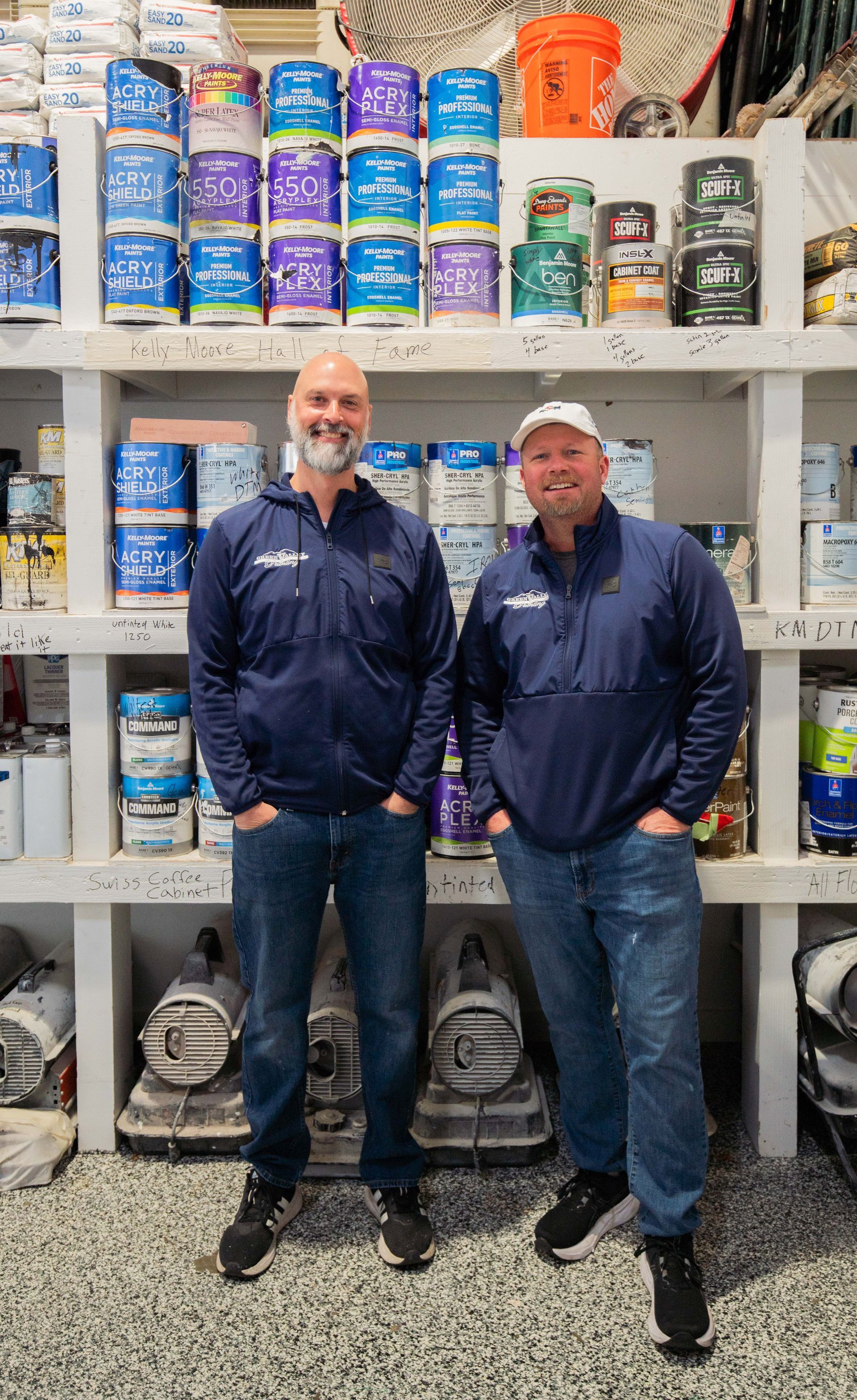 Two men are standing in front of a shelf full of paint cans.