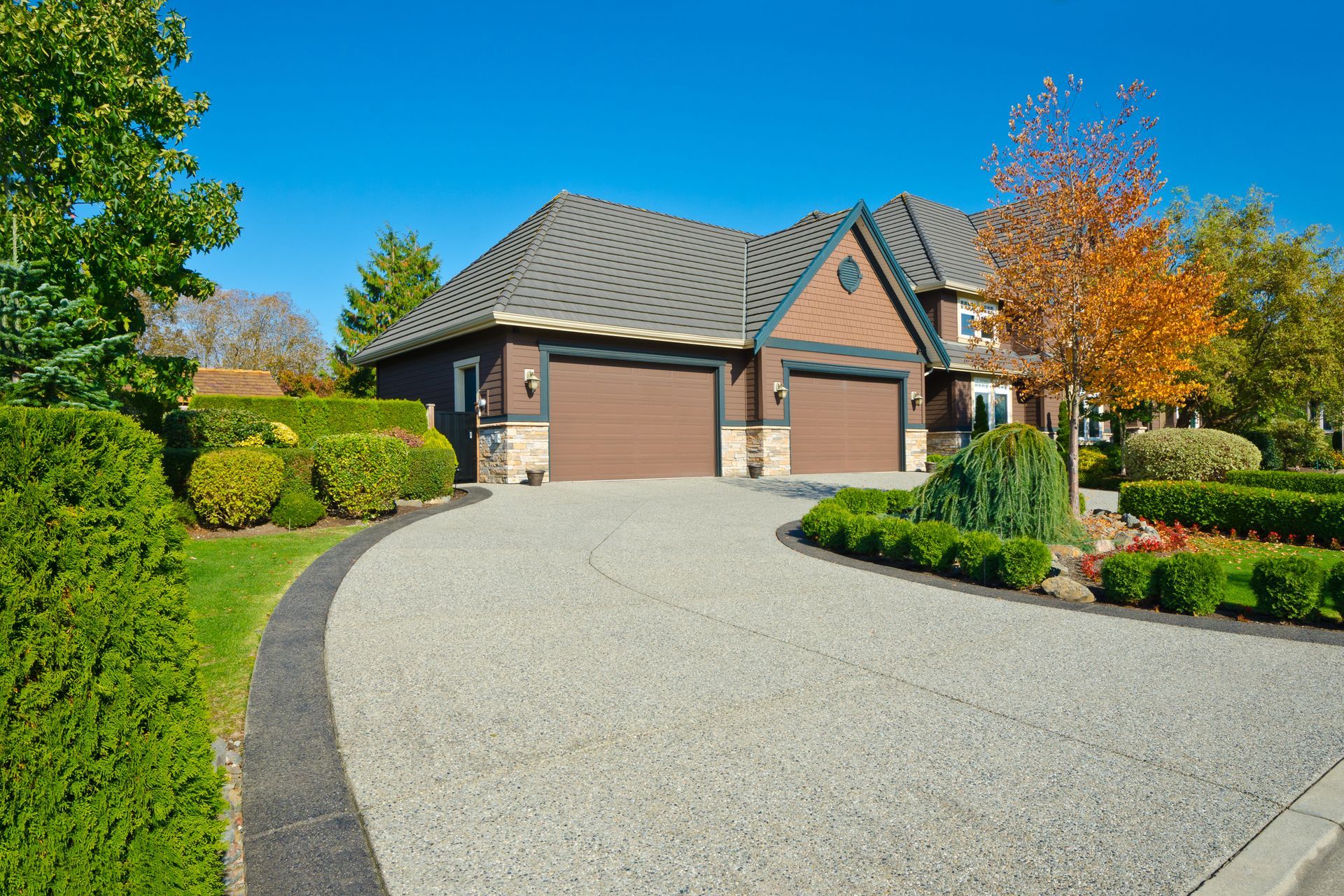 House with two-car garage, long driveway, and lush landscaping under a bright blue sky.