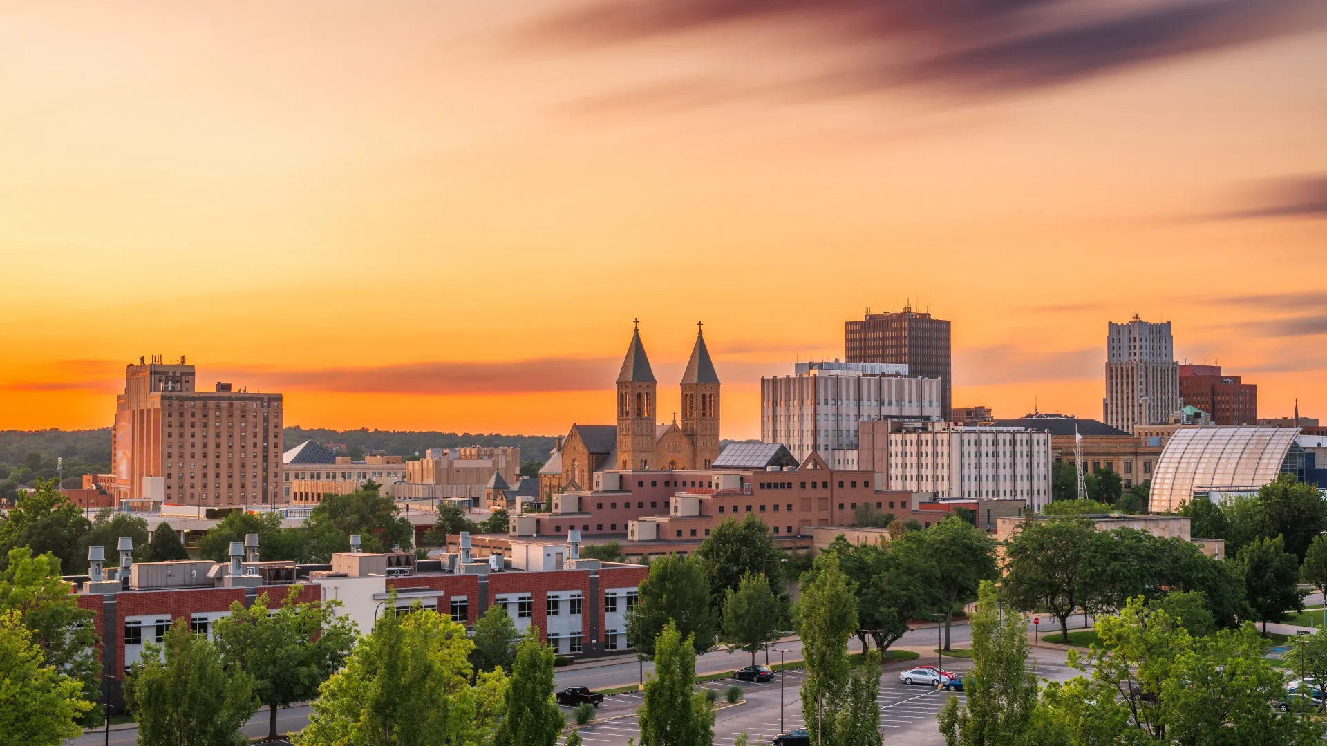 City skyline at sunset with a golden sky, featuring buildings, trees, and a church with two spires.