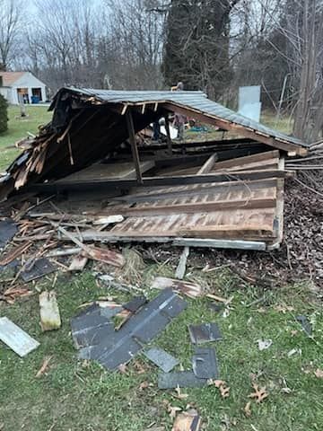 A partially collapsed wooden shed with scattered debris on a grassy yard.