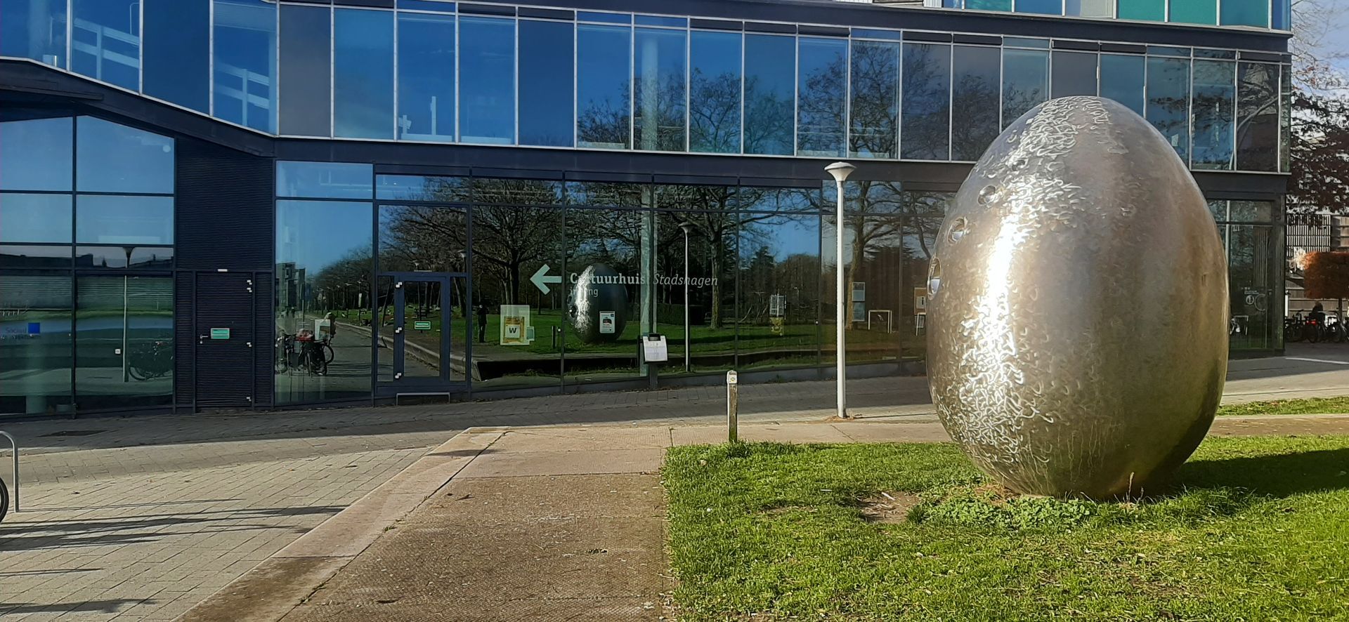 Een grote, metalen eivormige sculptuur voor een modern gebouw met reflecterende glazen ramen.