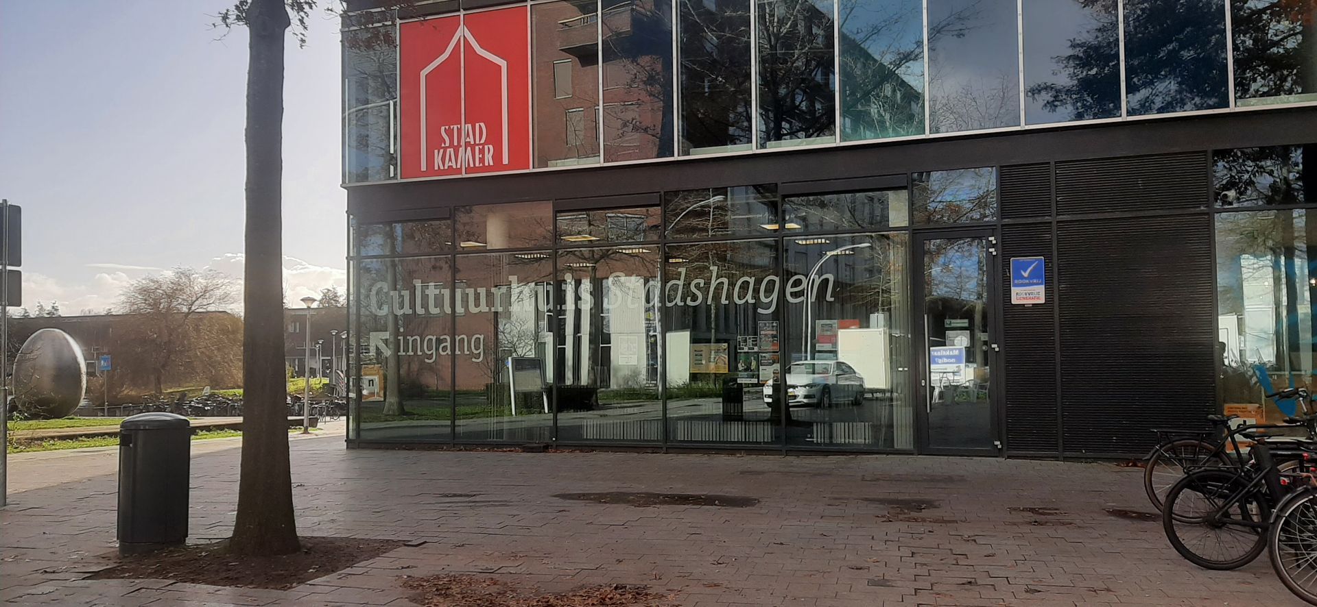 Een modern glazen gebouw met een rood uithangbord met een witte boog. Er is een trottoir en er staan ​​fietsen voor.