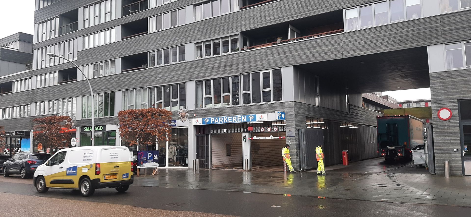 Een gele servicebus staat geparkeerd voor een gebouw met mensen in gele hesjes, mogelijk reparateurs.