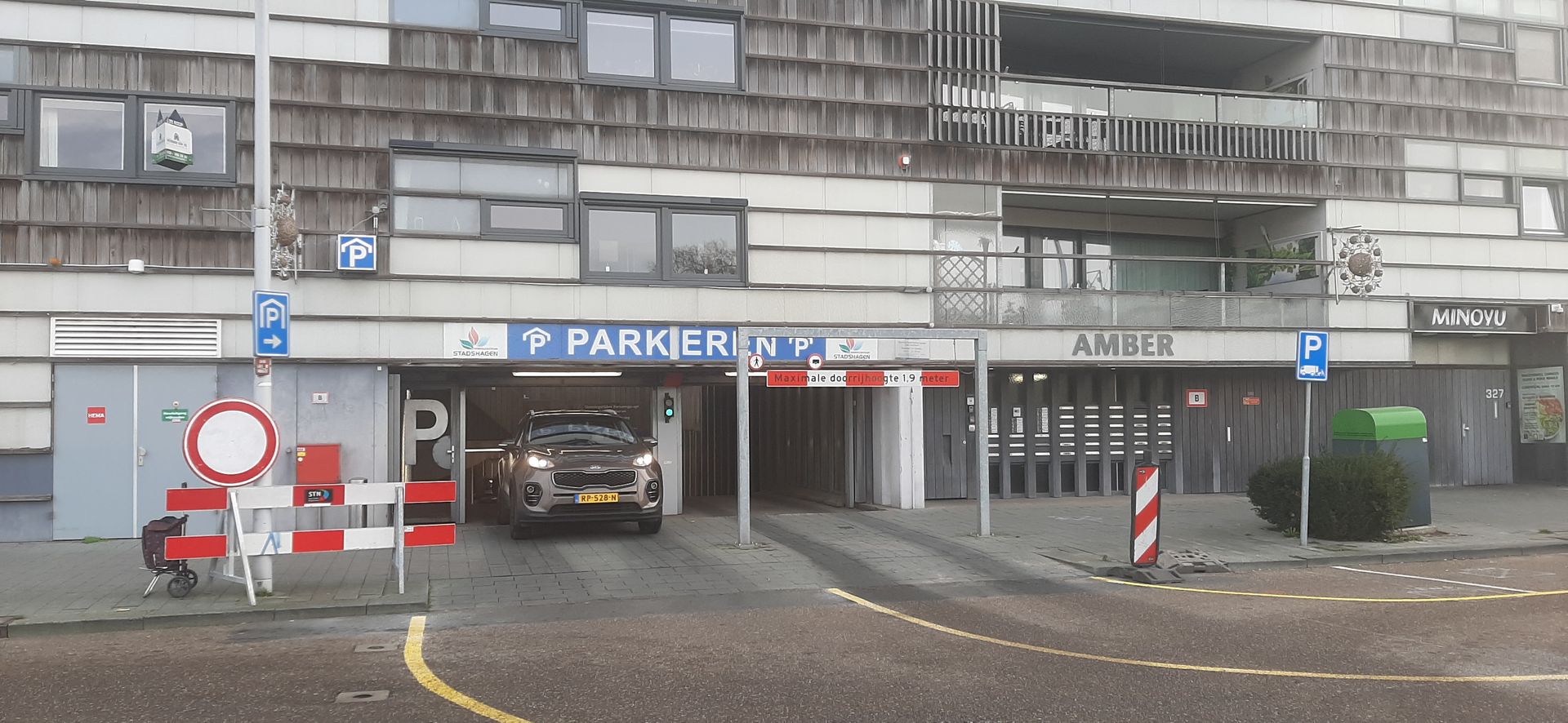 Ingang van een parkeergarage met een auto die naar binnen rijdt; rode en witte slagbomen en borden zijn op de voorgrond te zien.
