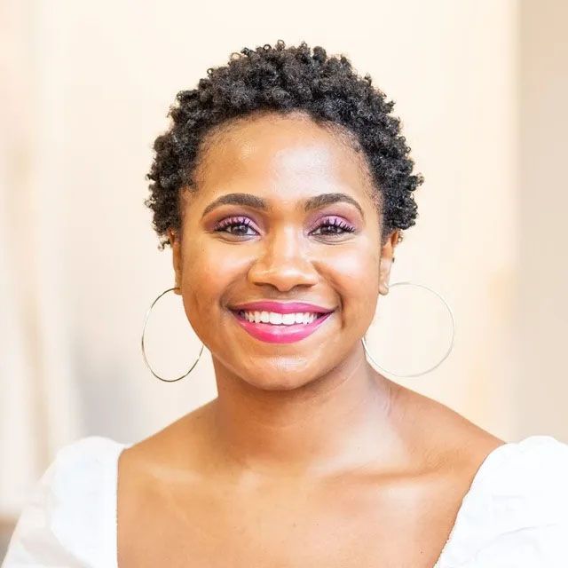A smiling person with short, curly hair and hoop earrings wearing a white top against a soft, neutral background.