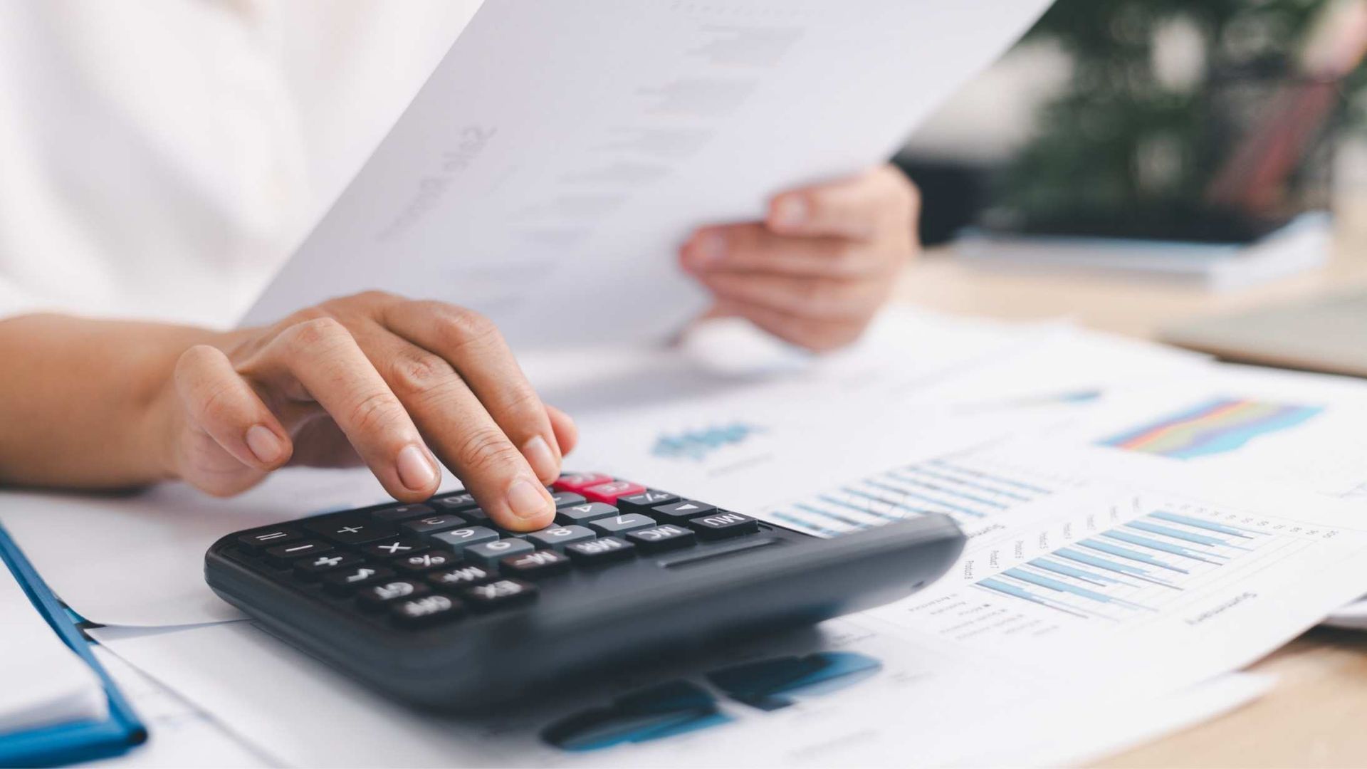 a person holding a document with their left hand and typing on a calculator with their right hand