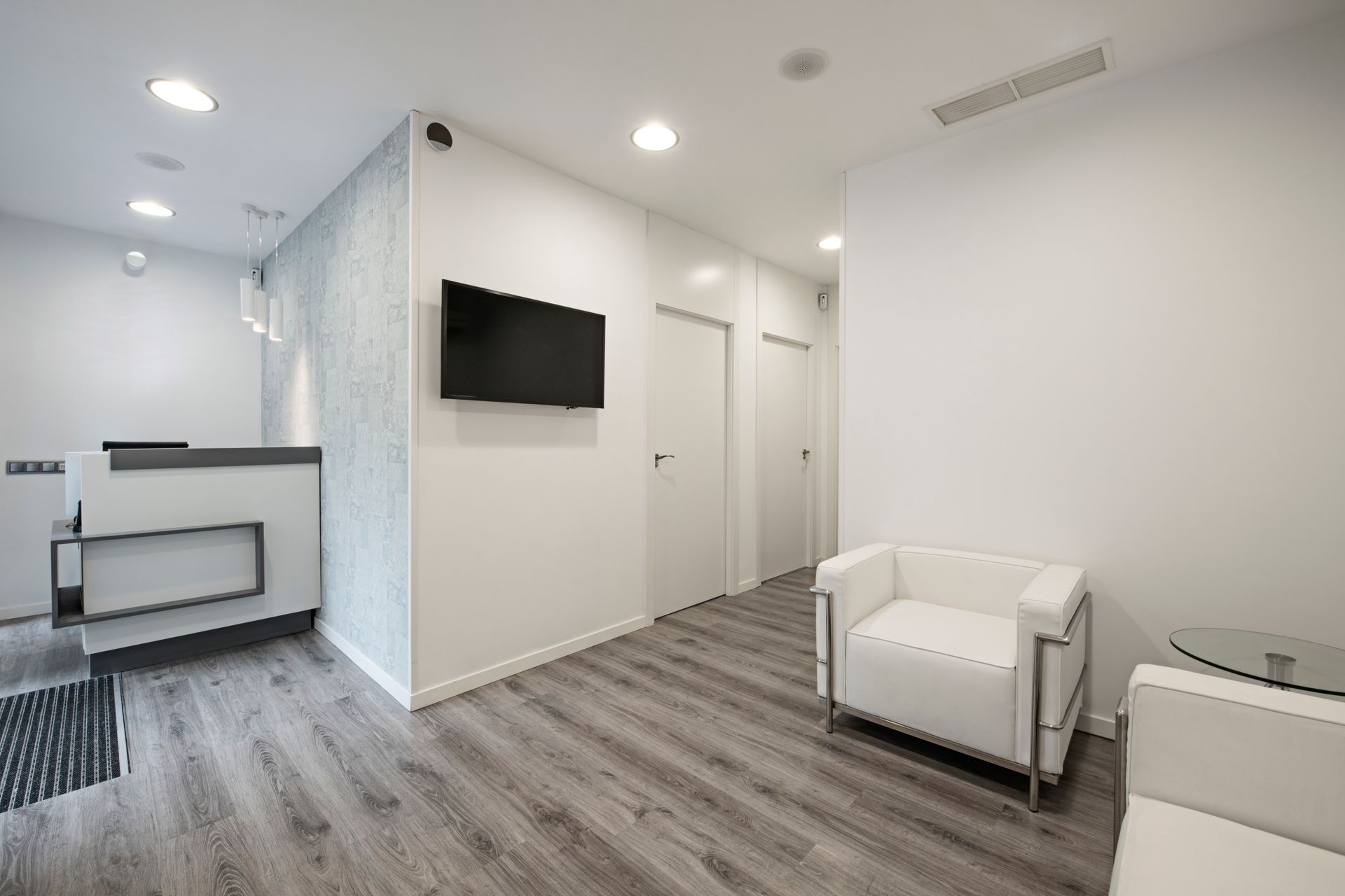 Salle d'attente minimaliste avec un comptoir d'accueil, une télévision murale, un sol gris imitation bois et des fauteuils blancs.