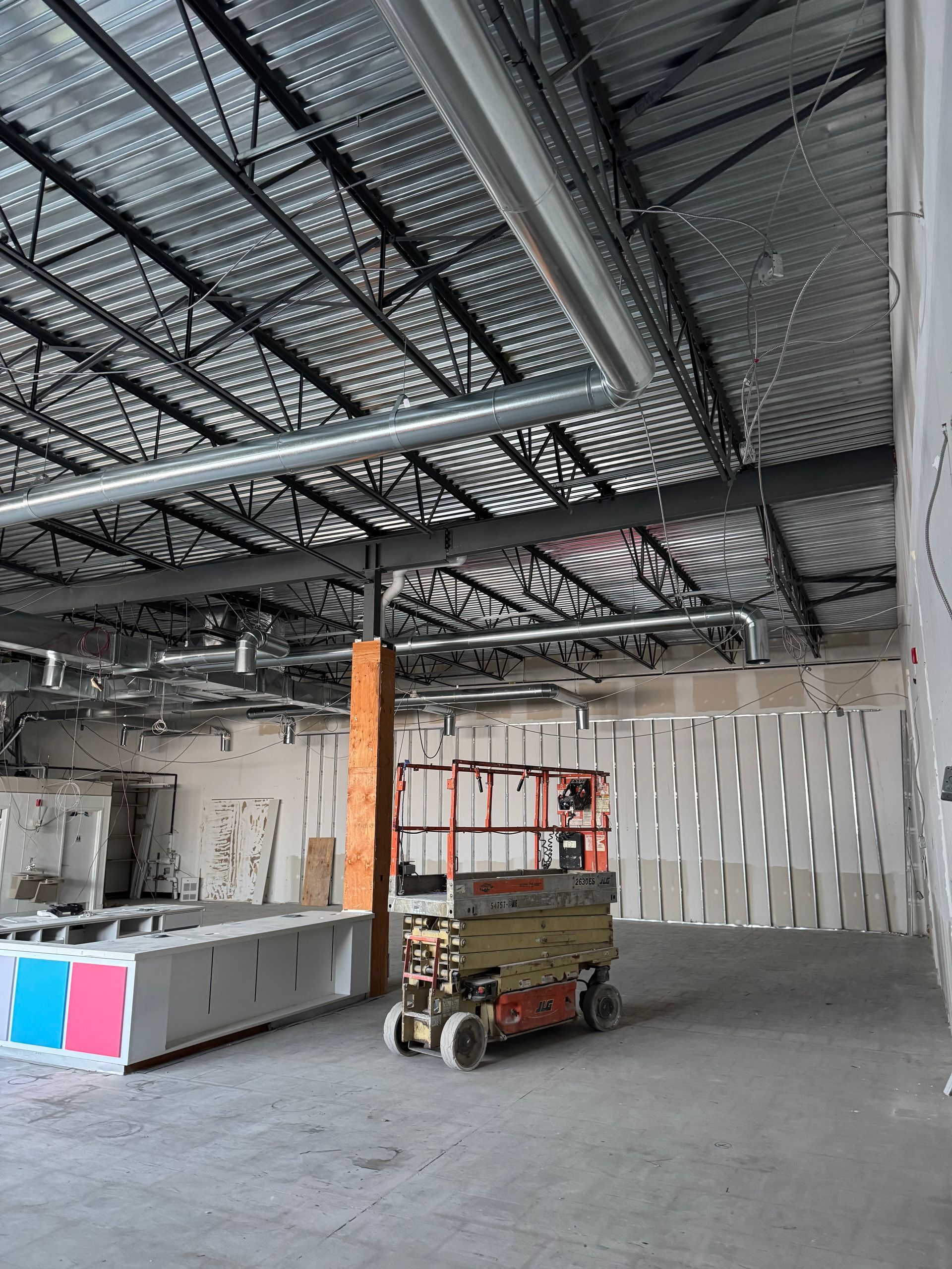 Un chantier de construction intérieur avec des plafonds apparents, des conduits de ventilation, une nacelle élévatrice au centre et une ossature métallique.