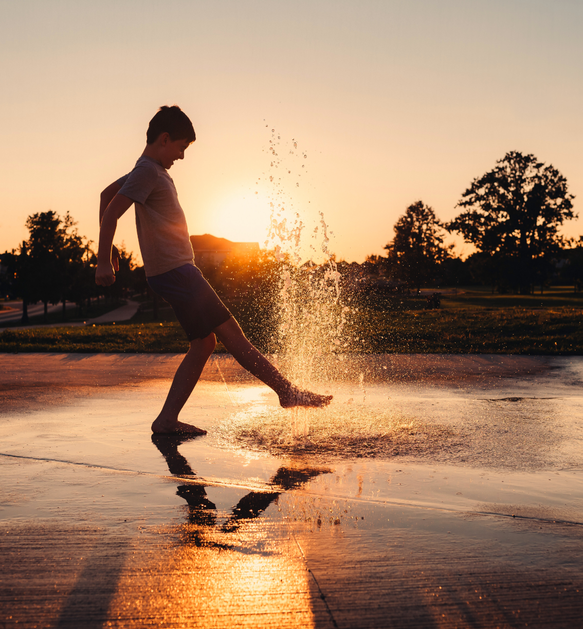 A young boy is kicking water in a puddle at sunset.