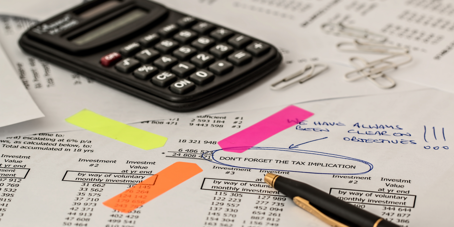 A black school calculator and a parker pen laying on top of paper spreadsheets which have writing and sticky notes on them 