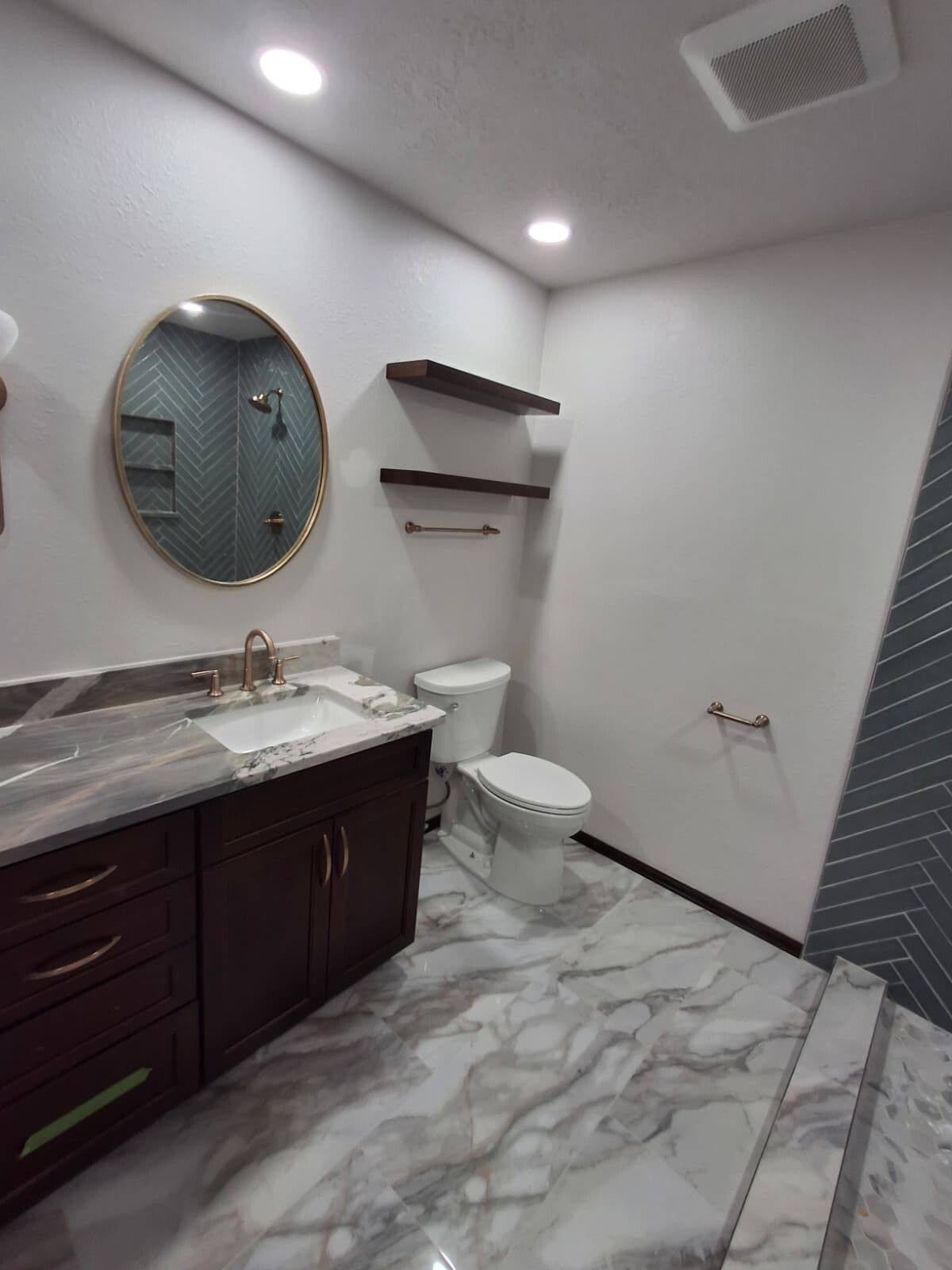 Small modern bathroom with white walls, marble floor, dark vanity, round mirror, toilet, and wall shelves