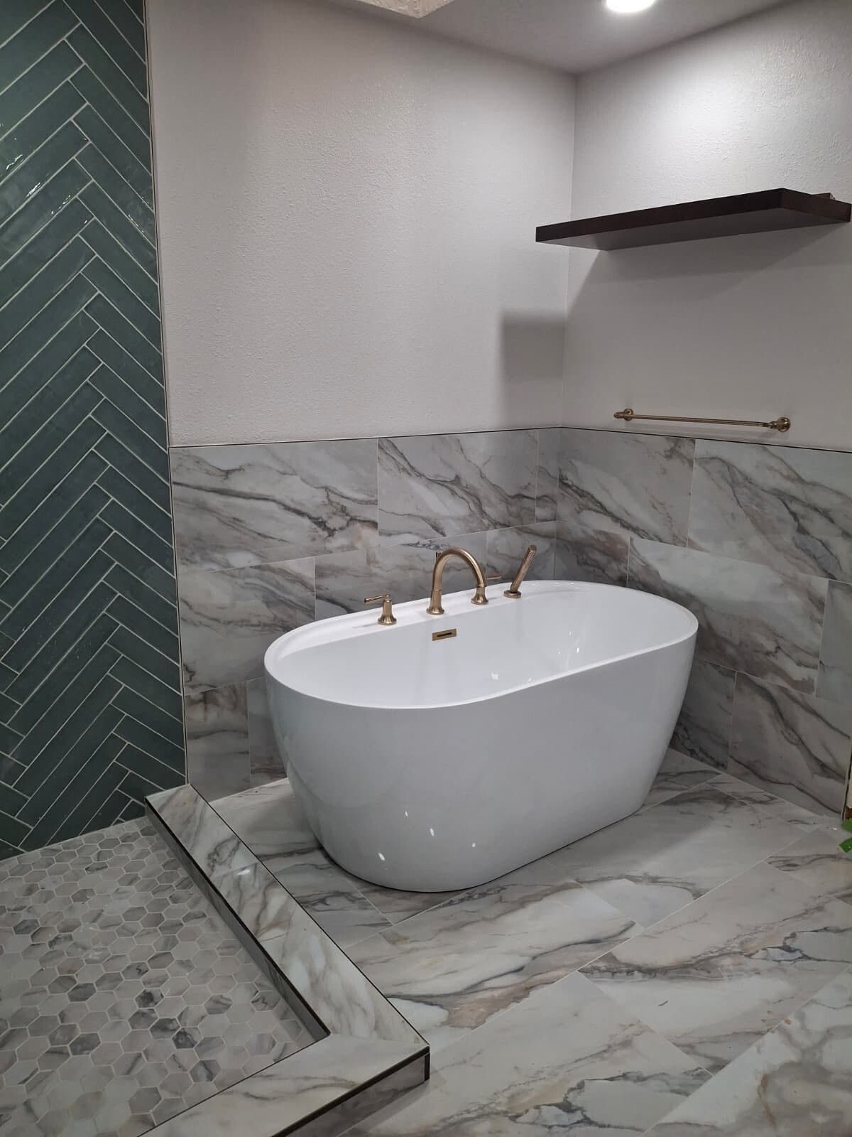 Modern bathroom with freestanding white tub, marble walls and floor, and dark green shower tile accent