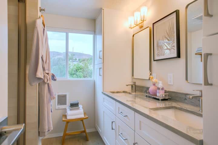 Bright bathroom vanity with white cabinets, mirror, and wall sconces beside a window and robe hook.