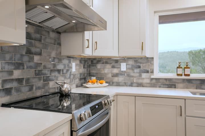Modern kitchen with white cabinets, gray tile backsplash, stainless steel stove, and a window with bottles on the sill