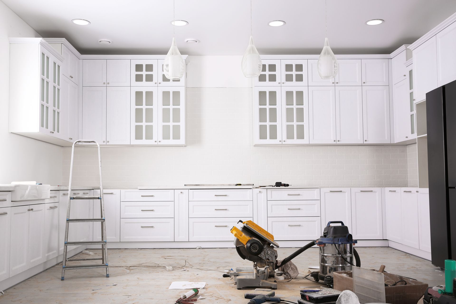 A kitchen mid-remodel with white shaker cabinets, pendant lights, a miter saw, and a vacuum cleaner A kitchen mid-remodel with white shaker cabinets, pendant lights, a miter saw, and a vacuum cleaner