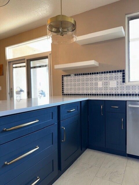 Blue kitchen cabinets with white countertops, mosaic backsplash, and pendant light in a bright modern room