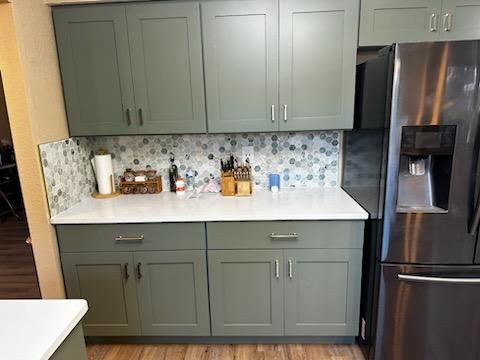 Modern kitchen with sage green cabinets, white counters, mosaic backsplash, and stainless steel refrigerator.