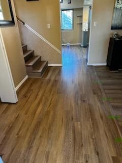 Empty hallway with wood floors, stairs on the left, and a doorway leading to a bright room.