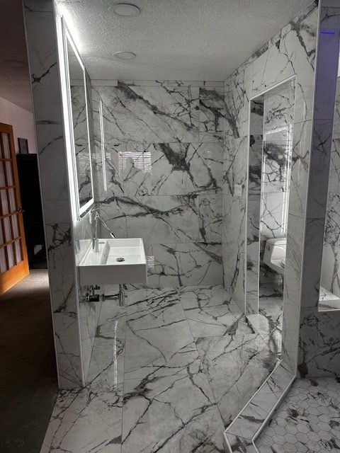 Marble-tiled shower with glass door, wall-mounted sink, and large mirror in a bathroom hallway