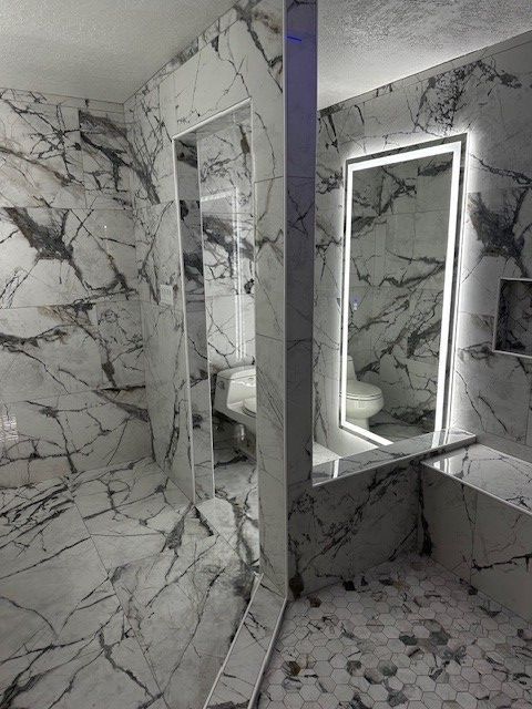 Marble-tiled bathroom with a lit vanity mirror, toilet, and walk-in shower with glass door.
