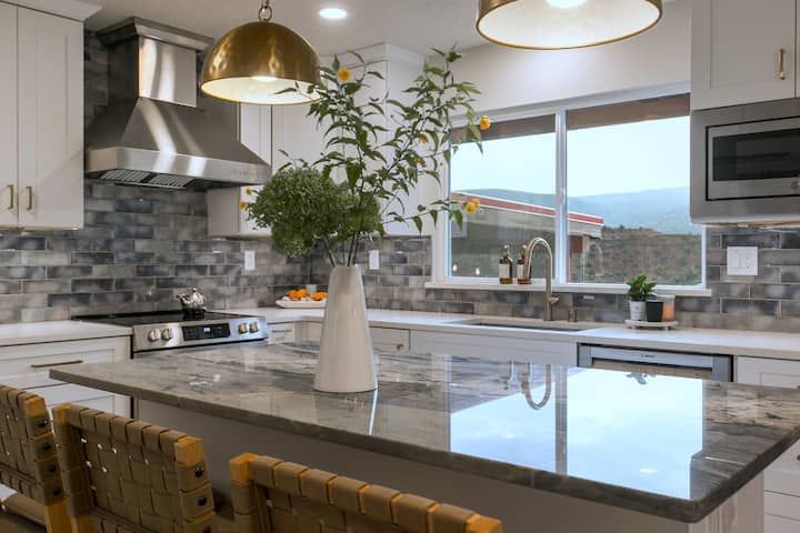 Modern kitchen with marble island, brass pendant lights, and a large window overlooking mountains.